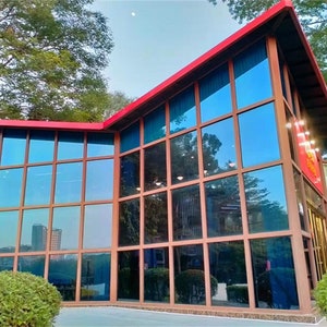 Puede incluir: Un edificio moderno con techo rojo y grandes ventanas tintadas de azul. El edificio tiene un dise&ntilde;o geom&eacute;trico con m&uacute;ltiples paneles de vidrio. El exterior est&aacute; enmarcado por madera marr&oacute;n. Arbustos verdes en primer plano y &aacute;rboles en el fondo.