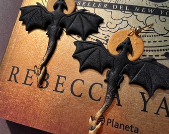 May include: A pair of black dragon earrings with gold accents. The dragons are flying with their wings spread out. The earrings are hanging from a book with the text "SANGRE" on the cover.