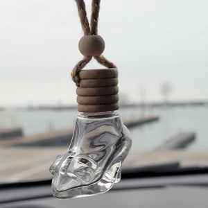 Puede incluir: Un ambientador de coche de vidrio transparente con forma de zapato de tac&oacute;n alto, colgado de un cord&oacute;n marr&oacute;n con cuentas de madera. El ambientador est&aacute; colgado del espejo retrovisor de un coche.