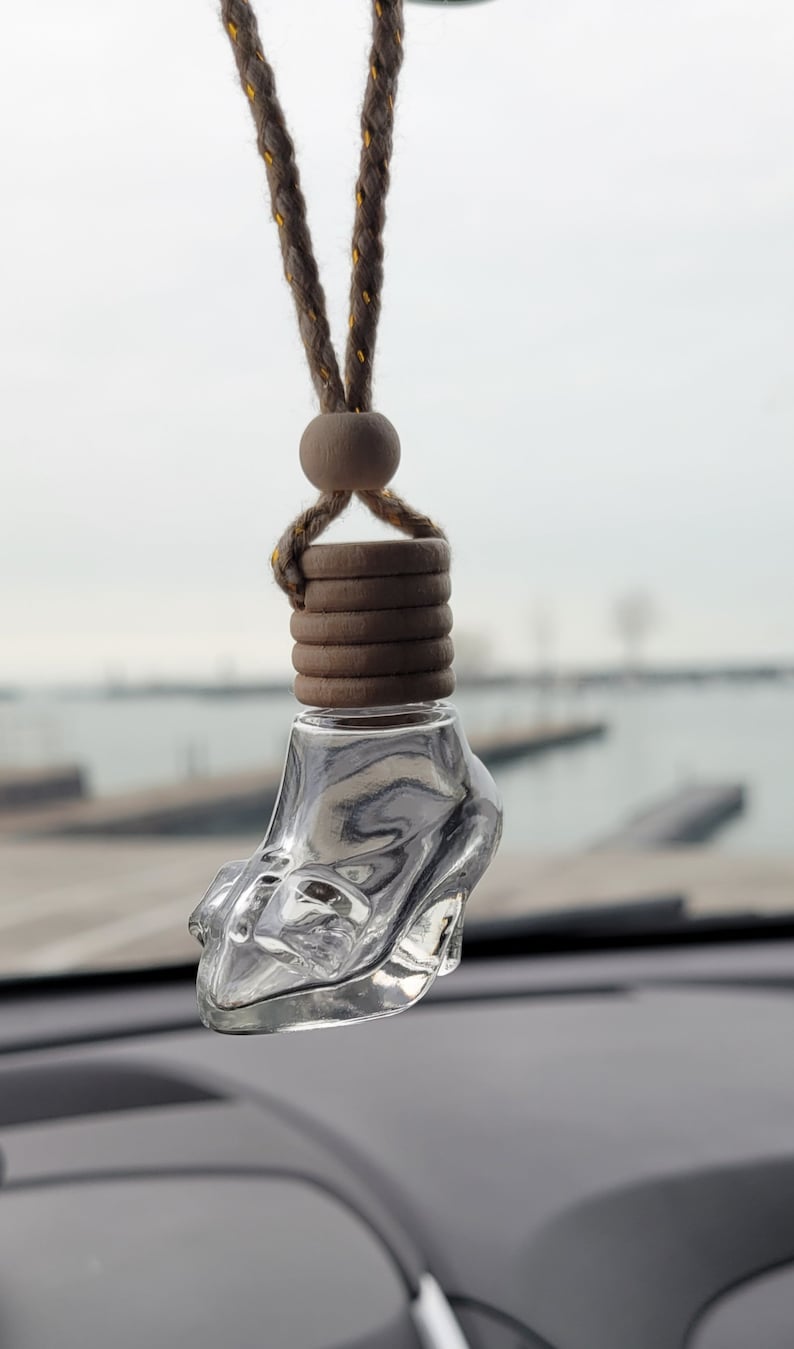 Puede incluir: Un ambientador de coche de vidrio transparente con forma de zapato de tac&oacute;n alto, colgado de un cord&oacute;n marr&oacute;n con cuentas de madera. El ambientador est&aacute; colgado del espejo retrovisor de un coche.