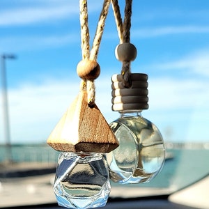 Puede incluir: Dos ambientadores de coche colgados de un espejo retrovisor. Uno es una botella de vidrio transparente con una tapa de madera y el otro es una botella de vidrio transparente con una tapa de madera geom&eacute;trica.
