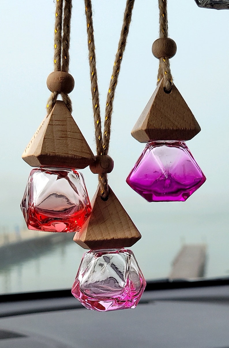 Puede incluir: Tres ambientadores de coche de madera con cuerdas colgantes. Cada ambientador tiene una botella de vidrio transparente con un l&iacute;quido rosa, rojo o morado en su interior. Las botellas tienen forma de diamante.