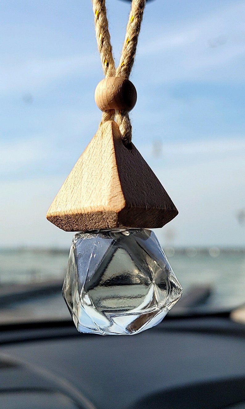 Puede incluir: Un ambientador de coche triangular de madera con una botella de vidrio transparente en forma de diamante que cuelga de una cuerda amarilla y blanca.