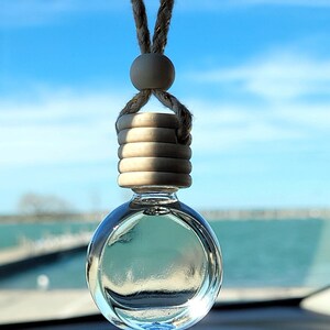 Puede incluir: Una botella colgante de vidrio transparente con una tapa de madera y una cuerda marr&oacute;n que cuelga del espejo retrovisor de un coche. La botella est&aacute; llena de un l&iacute;quido transparente y el fondo es un cielo azul y agua.