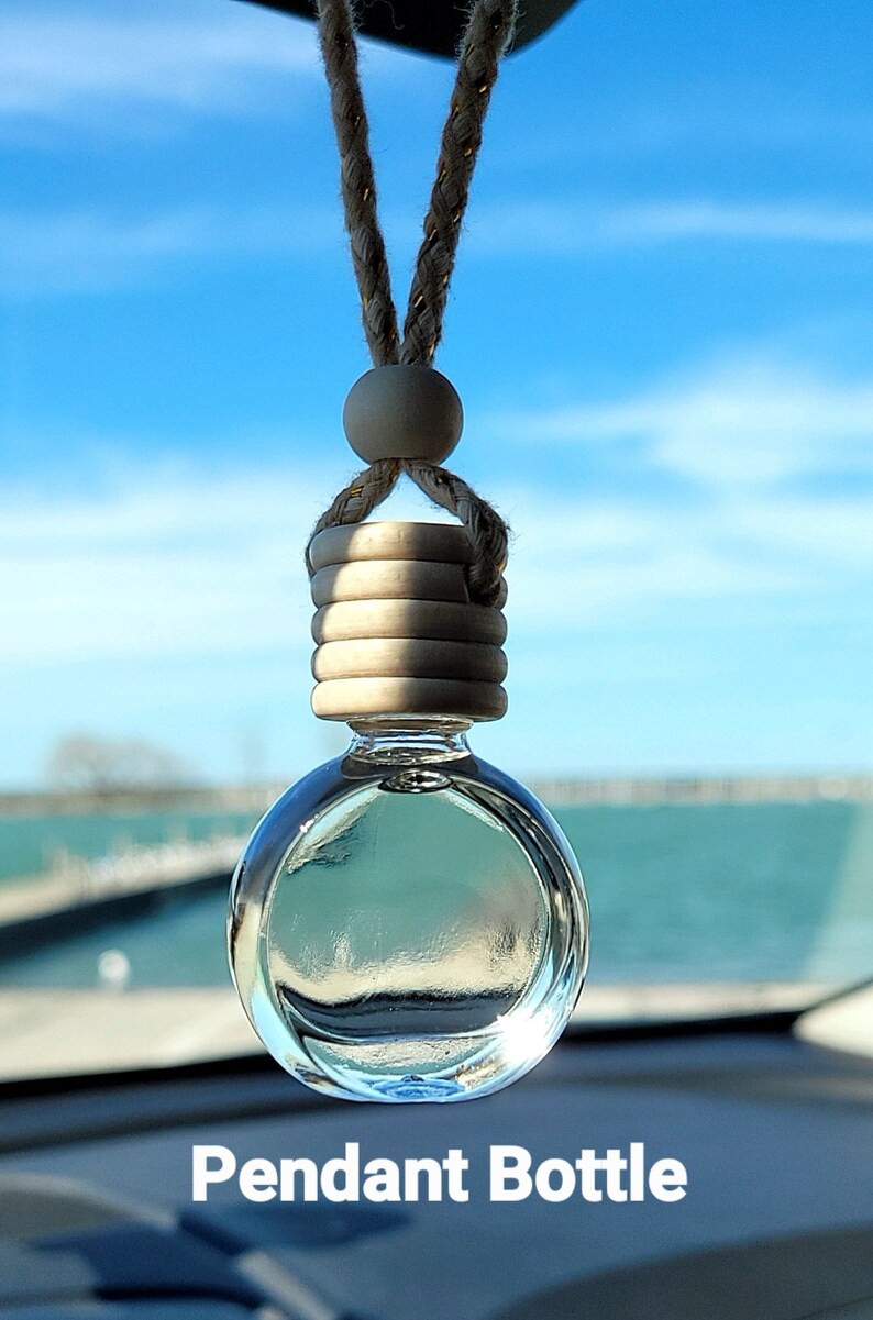 Puede incluir: Una botella colgante de vidrio transparente con una tapa de madera y una cuerda marr&oacute;n que cuelga del espejo retrovisor de un coche. La botella est&aacute; llena de un l&iacute;quido transparente y el fondo es un cielo azul y agua.
