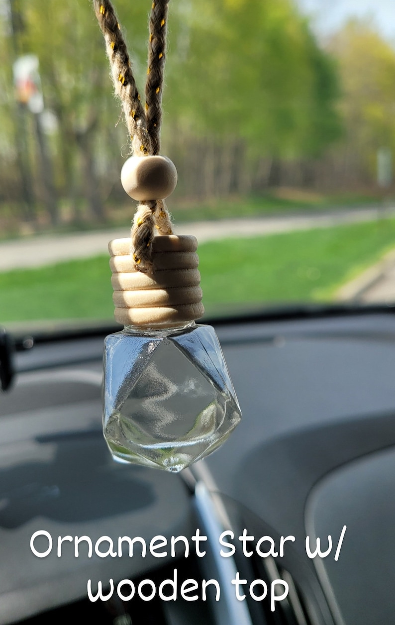 Puede incluir: Un adorno de vidrio transparente en forma de estrella con una parte superior de madera y una cuerda para colgar. El adorno est&aacute; colgado del espejo retrovisor de un coche.