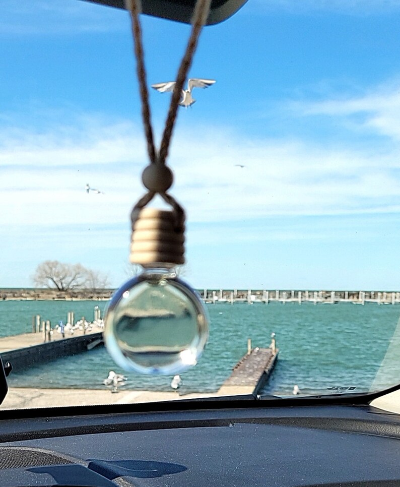 Puede incluir: Una botella de vidrio transparente con una tapa de madera cuelga de una cuerda en un coche. La botella est&aacute; llena de un l&iacute;quido transparente. La ventana del coche est&aacute; abierta y la vista exterior es de un cielo azul, nubes blancas y una masa de agua con un muelle en la distancia.