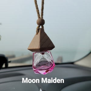 Puede incluir: Un ambientador de coche de vidrio rosa que cuelga de una cuerda marr&oacute;n con una parte superior de madera en forma de pir&aacute;mide. El texto "Moon Maiden" es visible debajo del ambientador.