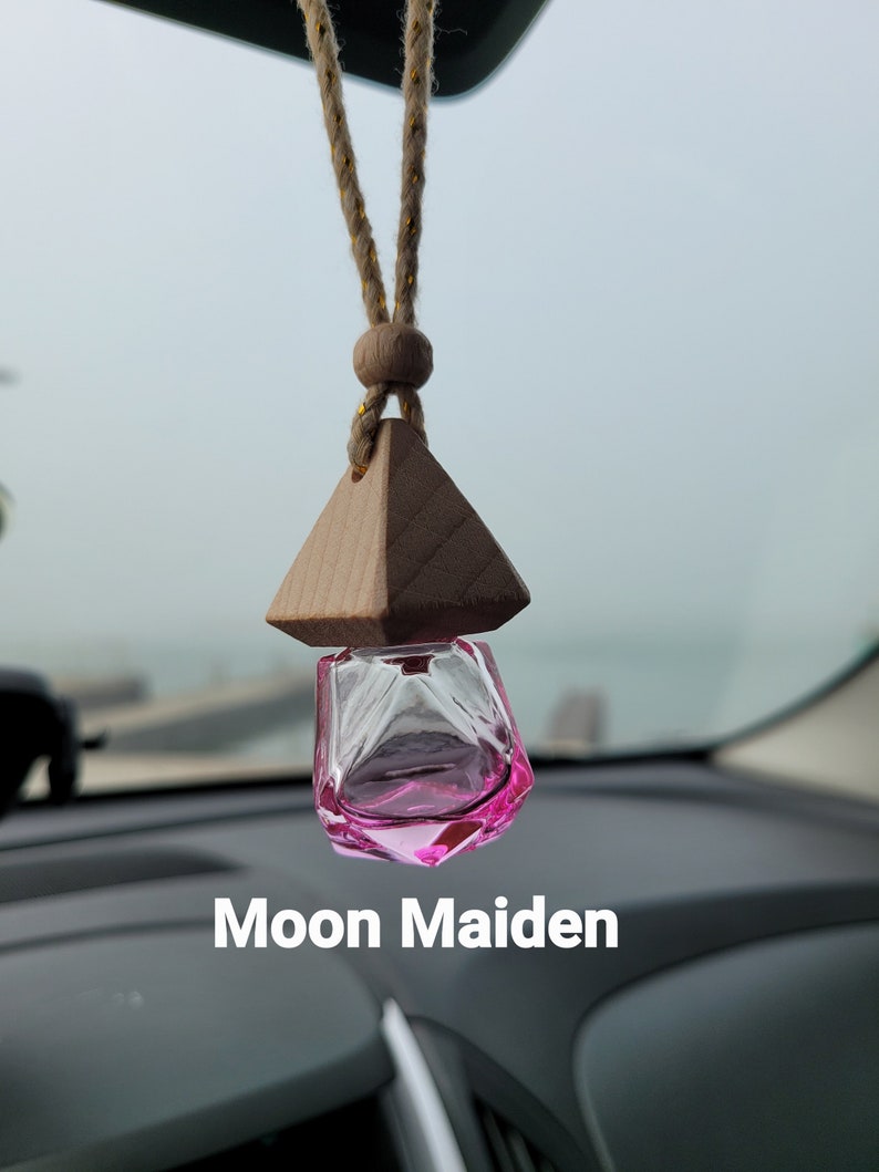 Puede incluir: Un ambientador de coche de vidrio rosa que cuelga de una cuerda marr&oacute;n con una parte superior de madera en forma de pir&aacute;mide. El texto "Moon Maiden" es visible debajo del ambientador.