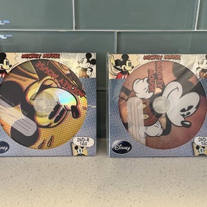 Puede incluir: Dos discos DVD-R de Mickey Mouse en su embalaje original. El embalaje presenta ilustraciones de Mickey Mouse y el logotipo de Disney. Los discos están etiquetados como "DVD-R 16X". El fondo es un azulejo azul claro.