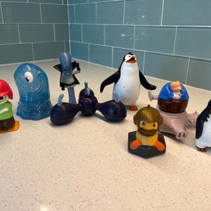 May include: A collection of colorful character figurines from the animated movie Madagascar. The figures include a penguin, a blue blob, and other characters, all standing on a white surface.