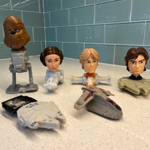 Puede incluir: Una colección de figuras y vehículos de personajes de Star Wars. El conjunto incluye a Chewbacca en un AT-ST, la Princesa Leia, Luke Skywalker, Han Solo, el Halcón Milenario y un caza estelar. Las figuras son de estilo caricatura.
