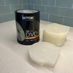 May include: A black TDK DVD-R container with a colorful disc graphic, stacked white disc cases, and a stack of clear DVD cases. The container reads "100PAQ DVD-R INscriptible 16X | 120MIN | 4.7GB".