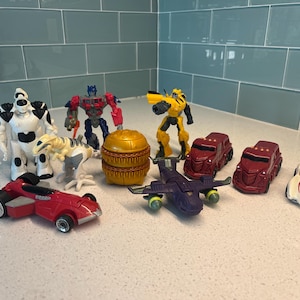 Può includere: Una collezione di personaggi e veicoli giocattolo colorati, tra cui robot, dinosauri e auto da corsa. I giocattoli sono di vari colori, come rosso, giallo, bianco e viola. I giocattoli sono disposti su una superficie bianca.