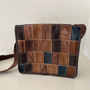 May include: A brown leather messenger bag with a patchwork design. The bag has a brown strap and is made of different pieces of leather with the words "Denim", "Canarc", and "Anarc" printed on them.