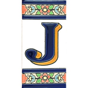 Könnte beinhalten: Eine Keramikfliese mit dem Buchstaben "J" in leuchtendem Blau mit orangefarbener Umrandung. Die Fliese wird von dekorativen Bändern mit floralen und geometrischen Mustern in Blau, Rot, Grün und Gelb umrahmt. Der Hintergrund ist weiß.