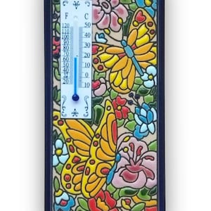 Può includere: Termometro decorativo con un design a farfalla e floreale. Il termometro mostra scale Celsius e Fahrenheit, con un indicatore di liquido blu. La cornice è blu scuro.