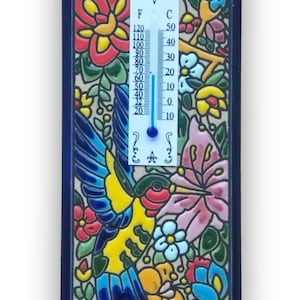 May include: A decorative ceramic thermometer with a vibrant floral and hummingbird design. The thermometer displays both Fahrenheit and Celsius scales. The artwork features a blue border and colorful flowers.