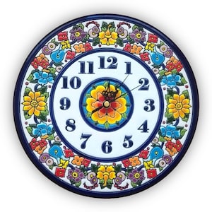 May include: Round ceramic wall clock with a white face and a blue border. The clock features black numerals and hands, and is decorated with colorful floral patterns in yellow, red, blue, and purple. The center of the clock has a large yellow flower.