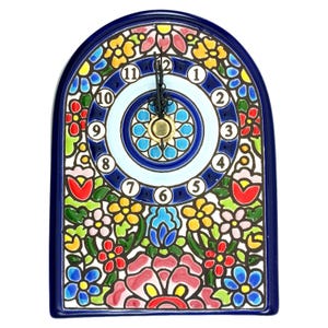Könnte beinhalten: Eine Keramikuhr mit einem dunkelblauen, bogenförmigen Rahmen. Das Zifferblatt ist von farbenfrohen Blumenmustern in Rot, Gelb, Blau und Grün umgeben. Die Zeiger der Uhr sind schwarz und die Zahlen weiß.