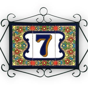 Può includere: Numero civico decorativo con il numero sette, caratterizzato da un vivace design floreale. Il numero è blu con un contorno dorato su sfondo bianco. La piastrella è incorniciata da un bordo in ferro battuto nero.