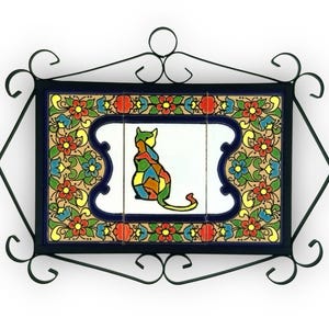 May include: A decorative tile featuring a colourful cat design. The cat is composed of various geometric shapes in multiple colours. The tile is surrounded by a floral pattern and framed in a black wrought iron border.