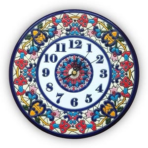 Puede incluir: Reloj de pared redondo de cerámica con esfera blanca y números azules. El reloj presenta un borde azul y un patrón floral decorativo en rojo, azul, amarillo y rosa. Las manecillas del reloj son negras.
