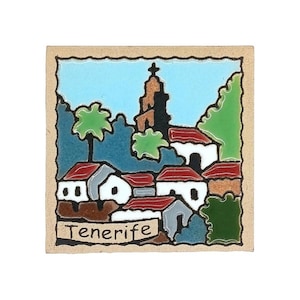 Op de afbeelding: Een vierkante keramische tegel met een handgeschilderde scène van een stad met witte gebouwen, rode daken, groene bomen en een hoge kerktoren tegen een lichtblauwe lucht. Het woord Tenerife staat in zwarte letters onderaan afgebeeld.