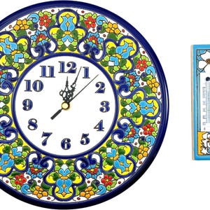 Könnte beinhalten: Eine runde Wanduhr aus Keramik mit weißem Zifferblatt, schwarzen Ziffern und einem dunkelblauen Rand mit einem bunten Blumenmuster in Gelb, Rot und Hellblau. Daneben befindet sich ein 15 cm hohes Thermometer aus Keramik.