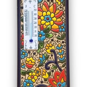 Op de afbeelding: Een decoratieve keramische thermometer met een bloemenontwerp. De thermometer geeft Fahrenheit- en Celsius-schalen weer. De achtergrond is beige met kleurrijke bloemen in rood, geel en blauw. De lijst is donkerblauw.