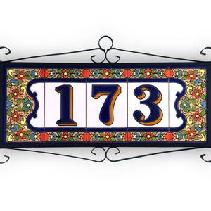 Può includere: Targa decorativa per indirizzo con il numero "173" in blu navy con dettagli arancioni su piastrelle bianche. Le piastrelle sono incorniciate da una cornice in ferro battuto nero, decorata con un motivo floreale.