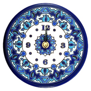 Op de afbeelding: Een ronde wandklok met een witte wijzerplaat en een blauw bloemmotief. De klok heeft een donkerblauwe rand en goudkleurige wijzers. De cijfers 12, 3, 6 en 9 zijn zichtbaar.