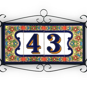 Pode incluir: Placa de endereço decorativa com o número "43" em azul marinho com detalhes em laranja sobre azulejos brancos. Os azulejos são emoldurados por uma borda floral em vermelho, amarelo, azul e verde. A placa é emoldurada por um design de ferro forjado preto.
