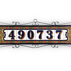 Könnte beinhalten: Dekorative Adressschild mit den Ziffern 490737 in Marineblau und Gold auf weißen Fliesen. Die Fliesen sind von einem schwarzen Schmiedeeisenrahmen mit dekorativen Schnörkeln und bunten floralen Akzenten umgeben.