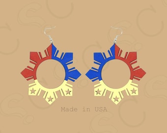 May include: A pair of dangling earrings featuring a sunburst design with eight rays. The earrings are colored in red, blue, and yellow, with three small stars at the bottom. Each earring hangs from a silver-toned metal hook. The text Made in USA is visible.