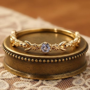 May include: A gold ring featuring a floral pattern and a small, pale blue gemstone at its centre. The ring is presented on a small, antique-style brass box, set against a lace background.