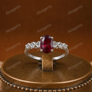 Puede incluir: Un anillo de tono plateado con una piedra preciosa de color rojo intenso y corte ovalado en el centro. La banda presenta un patrón geométrico de celosía texturizada a ambos lados de la piedra, creando un marco metálico detallado.