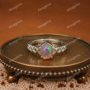 May include: A silver ring featuring an oval-shaped iridescent opal gemstone set in the center. The band of the ring has a decorative, open-work lattice pattern. The ring is positioned on a small, round, bronze-colored metal pedestal stand.