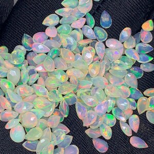 May include: A collection of iridescent opal gemstones in various shapes, including teardrops and ovals. The opals display a play of color with flashes of green, blue, pink, and yellow. The gemstones are scattered on a dark surface.