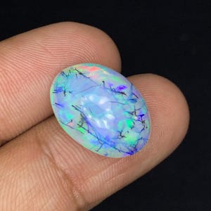 Puede incluir: Una piedra preciosa de ópalo de forma ovalada con una superficie translúcida e iridiscente. El ópalo muestra una mezcla de colores, incluyendo azul, verde y toques de rojo, con inclusiones oscuras que crean un patrón único. La gema se sostiene sobre un fondo oscuro.