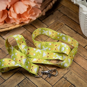 May include: A light green lanyard with a repeating pattern of yellow cartoon bunnies and white flowers. The lanyard has a silver-colored metal clasp. The background is a wooden surface.