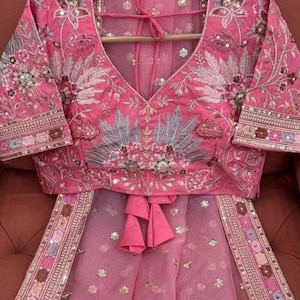 May include: A pink embroidered blouse with a V-neckline and short sleeves. The blouse is decorated with floral and leaf patterns in shades of pink, white, and grey. The blouse is paired with a sheer pink skirt with a floral pattern and a matching embroidered border.