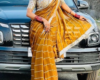 May include: A woman wearing a gold and white saree with a silver and white embroidered blouse. The saree has a white lace trim and is draped over a black car.