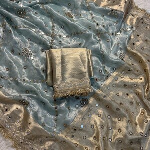 May include: A light blue and gold sari with intricate mirror and embroidery details. The sari features a shimmering, textured fabric with a gold border and matching gold fabric folded on top.