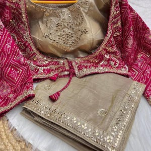 Peut inclure: Un sari en soie beige avec une bordure brodée d'or et un chemisier assorti brodé de rose et d'or. Le chemisier a un design à nouer au niveau du cou.