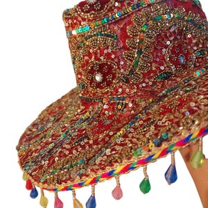 May include: A red, ornate sun visor with gold embroidery, sequins, and colorful beaded fringe. The visor features intricate patterns and embellishments, creating a festive and decorative accessory. The brim is trimmed with a rainbow-colored braid.