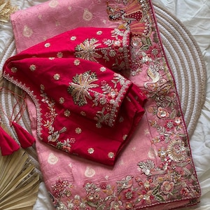 May include: A pink saree with intricate floral and bird embroidery, paired with a vibrant red blouse. The saree features detailed embellishments along the border and a matching embroidered blouse. The ensemble is displayed on a white surface.
