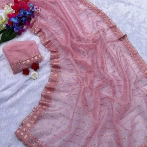 May include: A pink saree with gold detailing and a matching blouse. The saree is made of a sheer fabric with gold stripes and a decorative border. A bouquet of colorful flowers adds a vibrant touch to the scene.