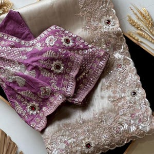 Puede incluir: Un sari bordado de lujo en tonos beige y morado. El sari presenta intrincados patrones florales y geométricos, adornados con lentejuelas y piedras preciosas rojas. La blusa morada tiene bordados detallados en las mangas y el escote.