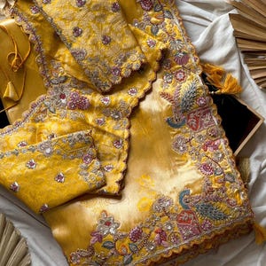 May include: A golden yellow sari with detailed embroidery. The sari showcases floral and peacock designs in pink, blue, and green. The edges are scalloped and decorated with sequins and tassels. The blouse piece is included.